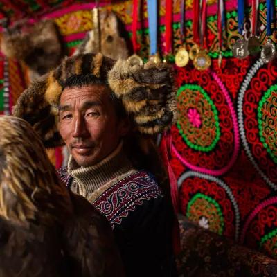 一个蒙古人站在一个明亮的蒙古包里，胳膊上挂着一只金鹰. 他戴着一顶毛皮帽子. 圆顶帐篷的墙上绣满了勋章.
