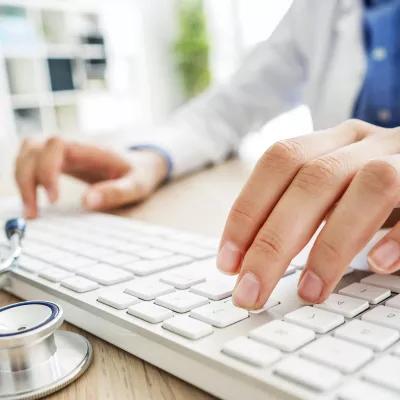 一个医疗专业人员在键盘上打字的特写，旁边放着听诊器.